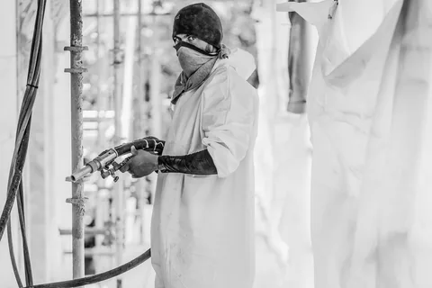 Industrial worker in a protective suit and mask spraying with a high-pressure gun on scaffolding